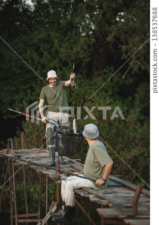 Father and adult son with fishing rods near the lake fishing at summer. Fishermen sitting on a footbridge. Men wearing khaki clothes and a caps. Father and adult son with fishing rods near the lake fishing at summer. Fishermen sitting on a footbridge. Men wearing khaki clothes and a caps. 138537688