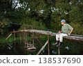 Man with fishing rod near the lake fishing at summer. Fisherman sitting on a footbridge. Man wearing khaki clothes and a cap. 138537690