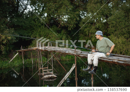 Man with fishing rod near the lake fishing at summer. Fisherman sitting on a footbridge. Man wearing khaki clothes and a cap. 138537690