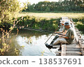 Two men with fishing rods near the lake fishing at summer. Fishermen sitting on a footbridge. Men wearing khaki clothes and a caps. 138537691