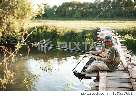 Two men with fishing rods near the lake fishing at summer. Fishermen sitting on a footbridge. Men wearing khaki clothes and a caps. 138537691