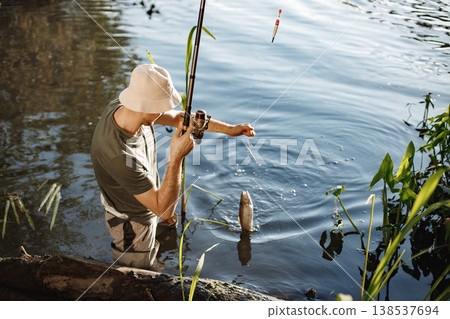 Man with fishing rod near the lake fishing at summer. Fisherman enjoying his hobby. Man wearing khaki clothes and a cap. Man with fishing rod near the lake fishing at summer. Fisherman enjoying his hobby. Man wearing khaki clothes and a cap. 138537694