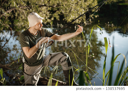 Man with fishing rod near the lake fishing at summer. Fisherman enjoying his hobby. Man wearing khaki clothes and a cap. 138537695