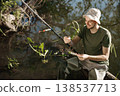 Man with fishing rod near the lake fishing at summer. Fisherman enjoying his hobby. Man wearing khaki clothes and a cap. 138537713