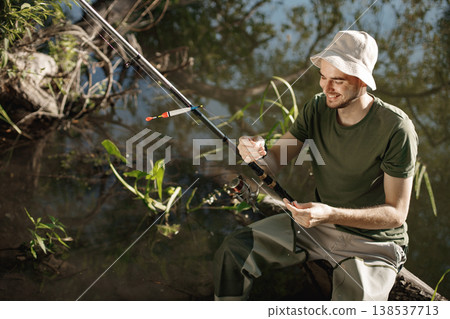 Man with fishing rod near the lake fishing at summer. Fisherman enjoying his hobby. Man wearing khaki clothes and a cap. Man with fishing rod near the lake fishing at summer. Fisherman enjoying his hobby. Man wearing khaki clothes and a cap. 138537713