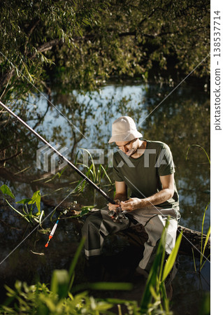 Man with fishing rod near the lake fishing at summer. Fisherman enjoying his hobby. Man wearing khaki clothes and a cap. Man with fishing rod near the lake fishing at summer. Fisherman enjoying his hobby. Man wearing khaki clothes and a cap. 138537714