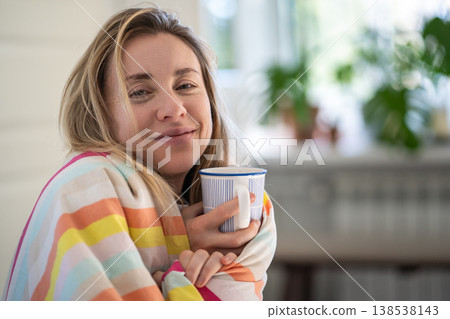 Dreamy woman wrapped in blanket enjoys slow morning and mug of aromatic coffee sitting in cozy bed. Dreamy woman wrapped in blanket enjoys slow morning and mug of aromatic coffee sitting in cozy bed. 138538143