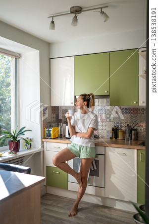 Cheerful woman standing in kitchen looking at window and enjoying morning coffee on weekend. Cheerful woman standing in kitchen looking at window and enjoying morning coffee on weekend. 138538191