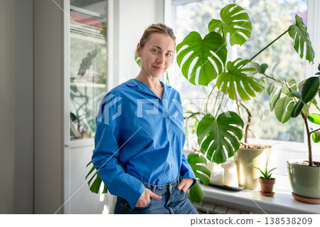Cheerful happy middle aged woman enjoying indoor gardening, posing near potted plants at windowsill. 138538209
