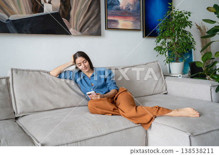 Smiling calm woman painter relaxing on couch procrastinating time scrolling media on smartphone. Smiling calm woman painter relaxing on couch procrastinating time scrolling media on smartphone. 138538211