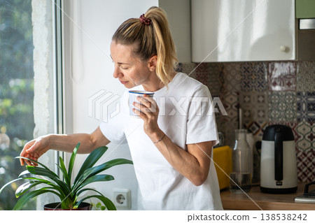 Positive woman with mug of coffee stands beside window at kitchen, slow morning at cozy home. Positive woman with mug of coffee stands beside window at kitchen, slow morning at cozy home. 138538242