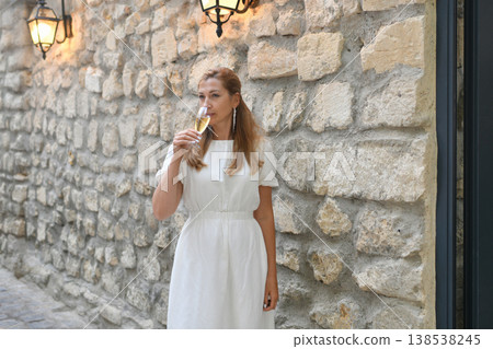 Woman sipping champagne against illuminated stone wall near venue door at a wedding Woman sipping champagne against illuminated stone wall near venue door at a wedding 138538245