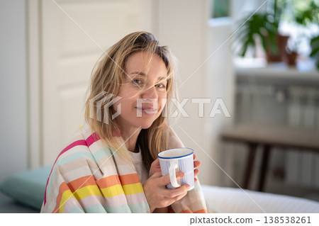 Cheerful tranquil woman with bed hair covered in blanket enjoys morning coffee sitting in comfy bed. 138538261