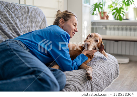 Cheerful mature woman pet lover comfortably lying and cuddling on sofa with beloved dog at home. Cheerful mature woman pet lover comfortably lying and cuddling on sofa with beloved dog at home. 138538304