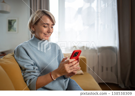 Beautiful middle aged woman sitting comfy on sofa and spending time scrolling on smartphone online. Beautiful middle aged woman sitting comfy on sofa and spending time scrolling on smartphone online. 138538309