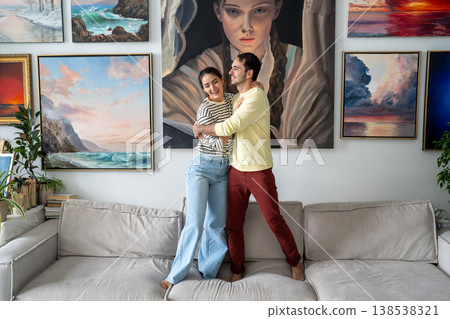 Happy married couple of painters man and woman hugging on sofa in front of home gallery exhibition. Happy married couple of painters man and woman hugging on sofa in front of home gallery exhibition. 138538321