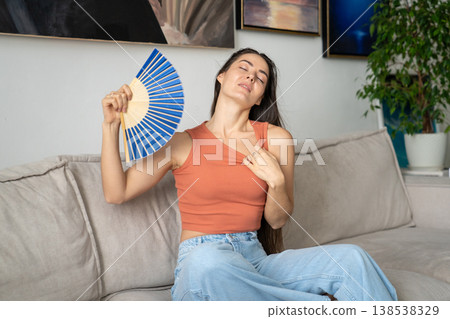 Carefree woman coping with summer heat, sits on sofa overwhelmed by heatwave, waving hand paper fan. Carefree woman coping with summer heat, sits on sofa overwhelmed by heatwave, waving hand paper fan. 138538329