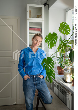 Cheerful happy middle aged woman enjoying indoor gardening, posing near potted plants at windowsill. 138538330