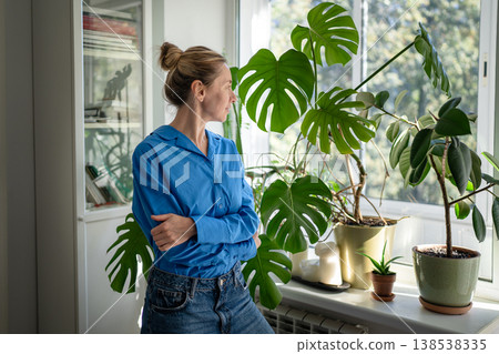 Thoughtful positive middle aged woman florist looking at own indoor garden on windowsill with love. Thoughtful positive middle aged woman florist looking at own indoor garden on windowsill with love. 138538335