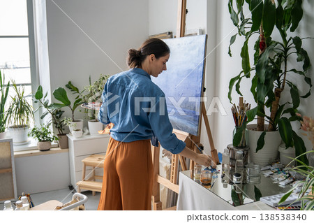 Thoughtful female painter enjoying hobby and drawing picture on easel at home studio apartment. Thoughtful female painter enjoying hobby and drawing picture on easel at home studio apartment. 138538340