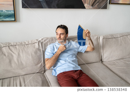 Guy coping with summer heat lying on sofa using paper fan suffocating of stuffiness, sweltering heat Guy coping with summer heat lying on sofa using paper fan suffocating of stuffiness, sweltering heat 138538348