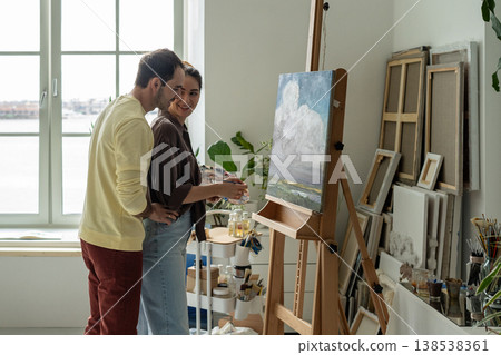 Cheerful woman artist painting on canvas in studio, husband enjoys process and supports wife painter Cheerful woman artist painting on canvas in studio, husband enjoys process and supports wife painter 138538361