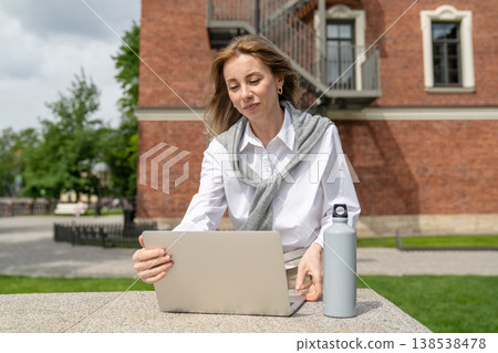 Cheerful businesswoman working on laptop outside in park, preparing for online business conference. Cheerful businesswoman working on laptop outside in park, preparing for online business conference. 138538478