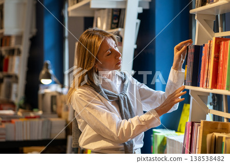 Intelligent woman booklover choosing books on rack filled with various publications in bookshop. Intelligent woman booklover choosing books on rack filled with various publications in bookshop. 138538482