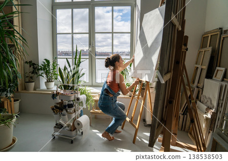 Concentrated female artist painting new artwork at loft workshop, working with canvas on easel. 138538503