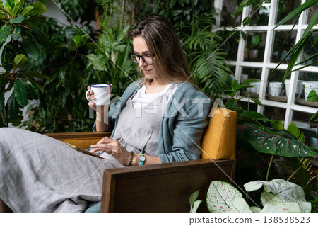 Woman gardener sitting on chair in home garden, resting, using smartphone and drinking tea from mug. Woman gardener sitting on chair in home garden, resting, using smartphone and drinking tea from mug. 138538523