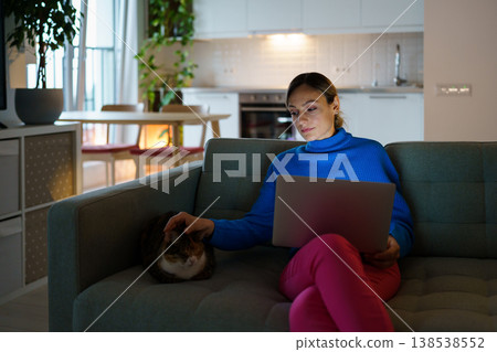 Woman freelancer sits on sofa in living room petting loving cat and watching webinar in laptop Woman freelancer sits on sofa in living room petting loving cat and watching webinar in laptop 138538552