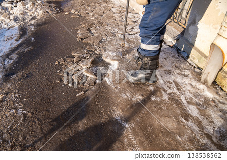 Street cleaner breaking ice with breaker tool, cleaning iced formation from drainpipe on sidewalk. Street cleaner breaking ice with breaker tool, cleaning iced formation from drainpipe on sidewalk. 138538562