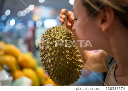 Woman cautiously sniffs durian on counter. Tropical exotic fruit choosing during grocery shopping Woman cautiously sniffs durian on counter. Tropical exotic fruit choosing during grocery shopping 138538574