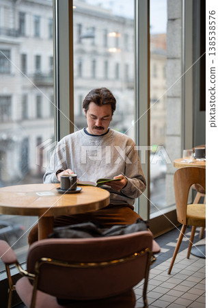 Man book lover fully engaged in reading popular book while sitting in cozy cafe with cup of coffee. Man book lover fully engaged in reading popular book while sitting in cozy cafe with cup of coffee. 138538576