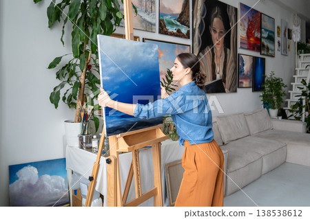 Focused woman artist working on painting in loft art gallery, adjusting finished artwork on easel. Focused woman artist working on painting in loft art gallery, adjusting finished artwork on easel. 138538612