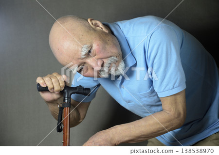 A bearded old man is doing rehabilitation indoors using a cane. 138538970