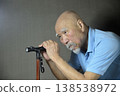 A bearded old man is doing rehabilitation indoors using a cane. 138538972