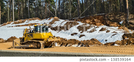 Yellow bulldozer pushing earth and grading a construction site in early spring, melting snow patches and bare trees visible at the forest edge under a clear blue sky. 138539164