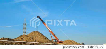 Construction crane working on a building site featuring large piles of earth and an electricity pylon against a clear blue sky, showing ongoing infrastructure development. Construction crane working on a building site featuring large piles of earth and an electricity pylon against a clear blue sky, showing ongoing infrastructure development. 138539177