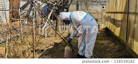 Person wearing a white protective suit, respirator mask, and gloves using a garden sprayer to apply chemicals to bare bushes and plants in a sunny garden for pest control and disease prevention. 138539182
