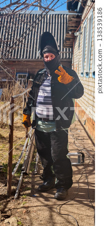 Welder in protective clothing with a welding mask raised, wearing work gloves and balaclava, smiling and showing a peace sign while standing outdoors next to a building. Welder in protective clothing with a welding mask raised, wearing work gloves and balaclava, smiling and showing a peace sign while standing outdoors next to a building. 138539186