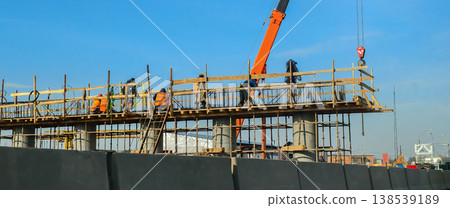 Construction workers wearing safety helmets and vests building a new highway overpass bridge, developing modern infrastructure with concrete pillars, formwork, scaffolding, and a crane. Construction workers wearing safety helmets and vests building a new highway overpass bridge, developing modern infrastructure with concrete pillars, formwork, scaffolding, and a crane. 138539189