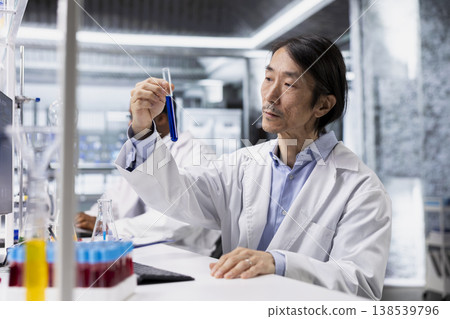 Old research facility employee using colorful chemical solutions in glassware. Asian chemical engineer in modern laboratory looking at blue reagent vial ready to be used for experimental analysis Old research facility employee using colorful chemical solutions in glassware. Asian chemical engineer in modern laboratory looking at blue reagent vial ready to be used for experimental analysis 138539796