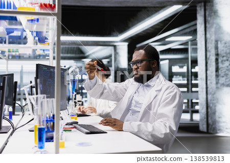 Quality control analyst in lab looking at blood sample in test tubes. African american man in high tech laboratory workspace analyzing sanguine fluid specimen in vial used for medical research Quality control analyst in lab looking at blood sample in test tubes. African american man in high tech laboratory workspace analyzing sanguine fluid specimen in vial used for medical research 138539813