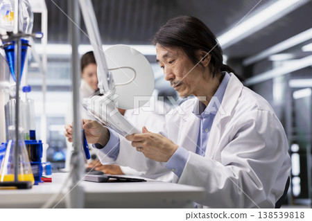 Molecular biologist uses laboratory lamp to examine liquid in test tube for clarity and color changes. Asian research facility expert holds test tube to light source for impurity detection 138539818