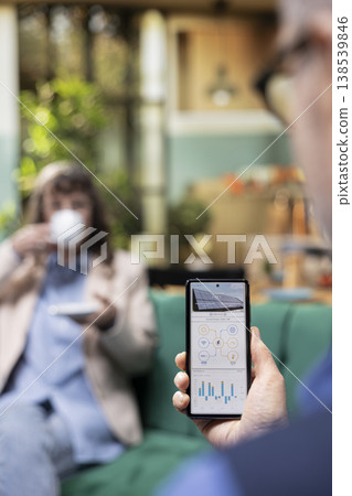 Senior people checking sustainable energy app on phone screen, relaxing together at home. Renewable solar power and photovoltaic panels highlight efficiency and environmental responsibility. Senior people checking sustainable energy app on phone screen, relaxing together at home. Renewable solar power and photovoltaic panels highlight efficiency and environmental responsibility. 138539846