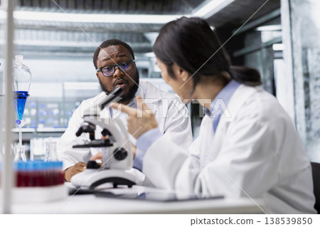 Diverse team of microscopists doing brainstorming in laboratory, inspecting specimen slide for microscopy study. Teamworking lab colleagues reviewing cytology slide for clinical testing procedure 138539850