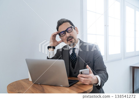A middle-aged male business professional struggling with a problem while using both a smartphone and a laptop. A middle-aged male business professional struggling with a problem while using both a smartphone and a laptop. 138541272