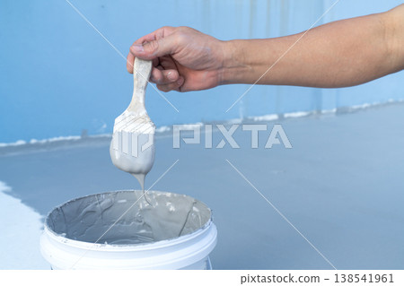 Hand holding a paintbrush dripping with gray paint over a white bucket. Professional home renovation and floor painting concept showing waterproof epoxy coating application on a blue surface. Hand holding a paintbrush dripping with gray paint over a white bucket. Professional home renovation and floor painting concept showing waterproof epoxy coating application on a blue surface. 138541961