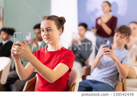 Group of teen listeners, girl take photos of presentation slide or scan QR code, teacher in background Group of teen listeners, girl take photos of presentation slide or scan QR code, teacher in background 138542439